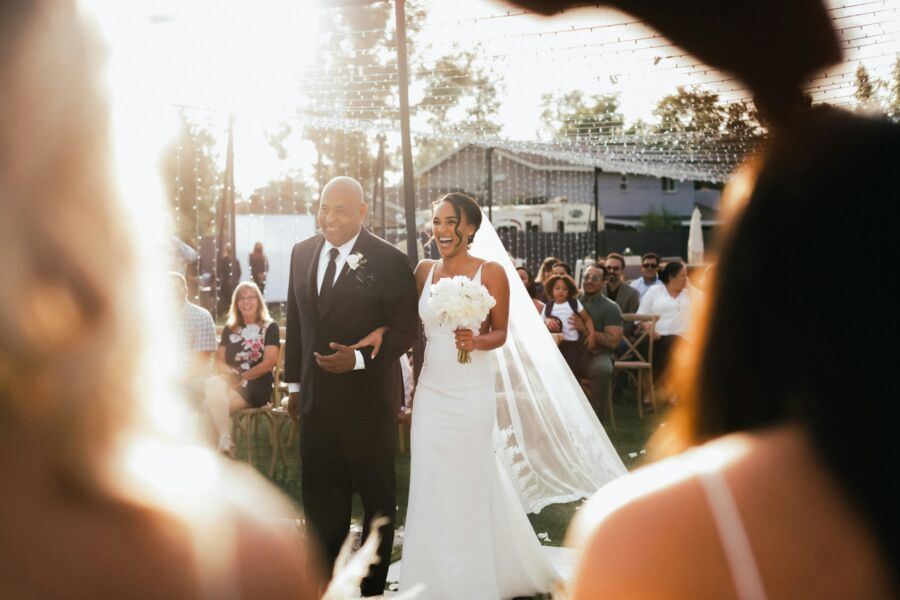 A bride walking the aisle arm in arm at golden hour, guests watching